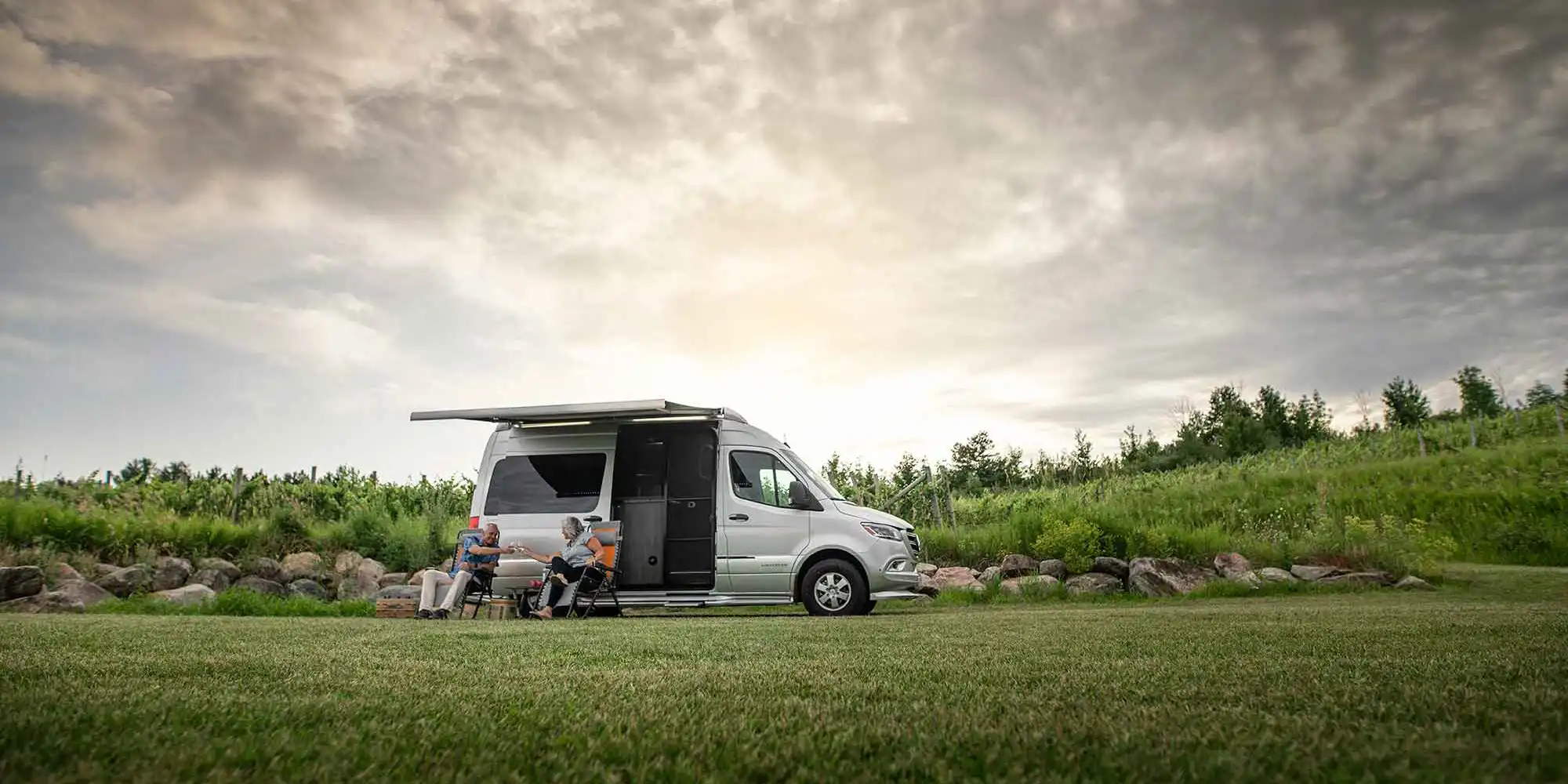 An Airstream Interstate touring coach beside a vineyard at dusk, two people relaxing in camp chairs